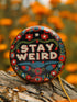 Stay Weird" black pinback button with pearl-effect lettering surrounded by folk art flowers in red and turquoise on a dark background.