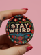 Stay Weird" black pinback button with pearl-effect lettering surrounded by folk art flowers in red and turquoise on a dark background.