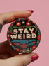 Stay Weird" black pinback button with pearl-effect lettering surrounded by folk art flowers in red and turquoise on a dark background.