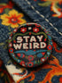Stay Weird" black pinback button with pearl-effect lettering surrounded by folk art flowers in red and turquoise on a dark background.