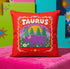 A square vibrant red throw pillow featuring a central illustration of the Taurus zodiac sign. The word TAURUS appears at the top in white bubbly letters with a faux-stitch texture. Below the text is an arch containing a purple bull standing in a green field dotted with white daisies and red flowers. Behind the bull are green pine trees and a large yellow sun against a blue sky. The design is framed by a pink wavy border, printed to look like raised embroidery on linen-textured polyester fabric.