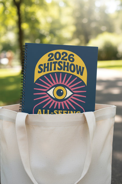 A hardcover spiral planner for 2026 with a dark blue cover and retro 70s style graphics. The title reads "2026 SHITSHOW All-Seeing Agenda" in bold yellow and pink text. Below the title is a large, stylized all-seeing eye with pink rays emanating from it. At the bottom, a pink ribbon contains the text "BLINK TWICE FOR MERCY." The back cover is also dark blue with a yellow sun graphic and the text "Plans bloom. Deadlines hum. I write, I wander, then get it done. Keep receipts. Keep grace.
