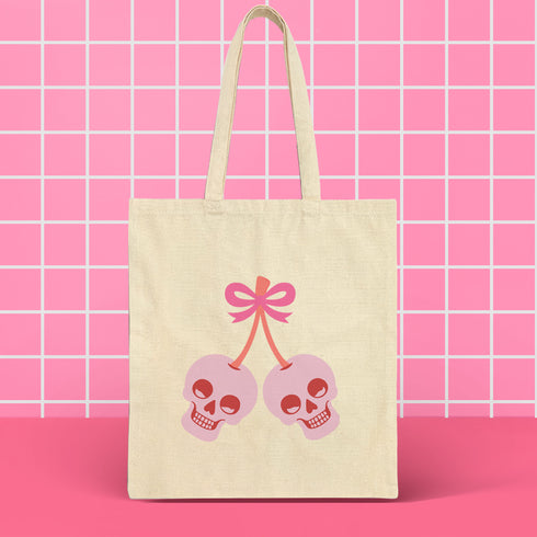 Canvas tote bag featuring pink skull cherries design. Two pink skulls connected by red stem and bow. Quirky gothic-inspired graphic on cream background. Stylish, edgy accessory for everyday use. Perfect for alternative fashion lovers.