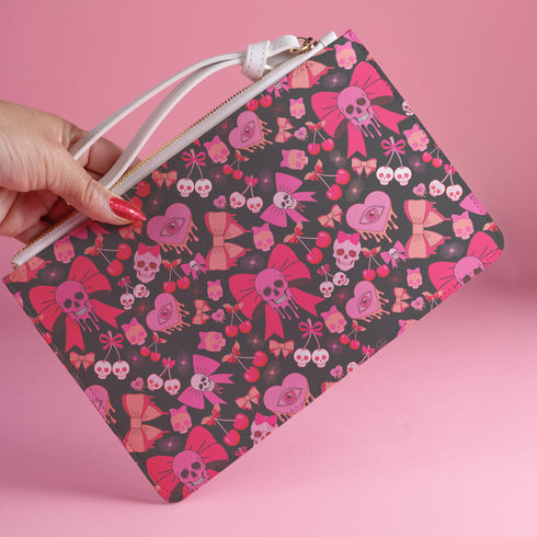 Rectangular wristlet clutch featuring a dark background with pink skull bows, cherries, and other goth-inspired motifs. White strap and gold zipper visible.
