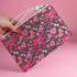 Rectangular wristlet clutch featuring a dark background with pink skull bows, cherries, and other goth-inspired motifs. White strap and gold zipper visible.