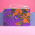 Rectangular wristlet clutch featuring vibrant purple and orange background with colorful snake prints. White strap and gold zipper visible.