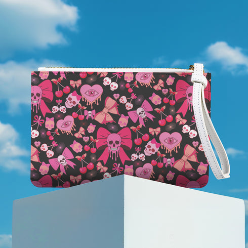 Rectangular wristlet clutch featuring a dark background with pink skull bows, cherries, and other goth-inspired motifs. White strap and gold zipper visible.