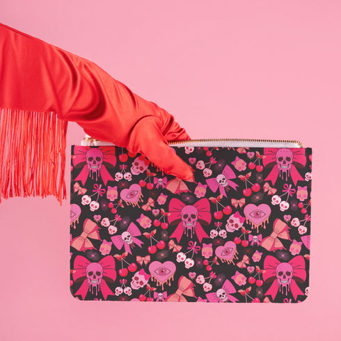 Rectangular wristlet clutch featuring a dark background with pink skull bows, cherries, and other goth-inspired motifs. White strap and gold zipper visible.
