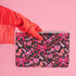 Rectangular wristlet clutch featuring a dark background with pink skull bows, cherries, and other goth-inspired motifs. White strap and gold zipper visible.