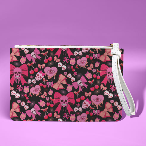 Rectangular wristlet clutch featuring a dark background with pink skull bows, cherries, and other goth-inspired motifs. White strap and gold zipper visible.