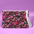 Rectangular wristlet clutch featuring a dark background with pink skull bows, cherries, and other goth-inspired motifs. White strap and gold zipper visible.