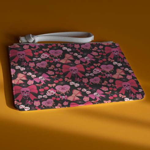 Rectangular wristlet clutch featuring a dark background with pink skull bows, cherries, and other goth-inspired motifs. White strap and gold zipper visible.
