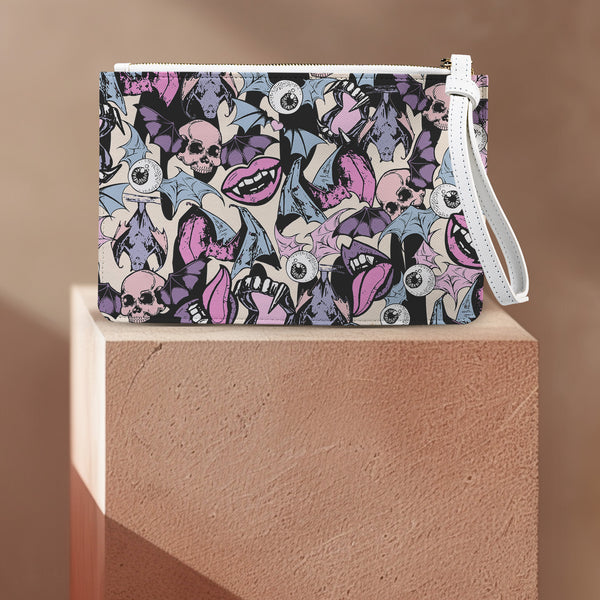 Rectangular wristlet clutch featuring a dark background with colorful bats, skulls, eyeballs, and vampire mouths. White strap and gold zipper visible.
