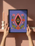 A vertical art print featuring a central pink yoni shape with a black interior containing a single eye. Golden yellow sun rays radiate from the center against a solid periwinkle blue purple background. The central motif is surrounded by a symmetrical botanical border of pink and red daisy flowers and green leaves, with two yellow crescent moons in the bottom corners. Printed on flat museum grade matte paper. This is the printed artwork only; a frame is not included. Ready to frame.