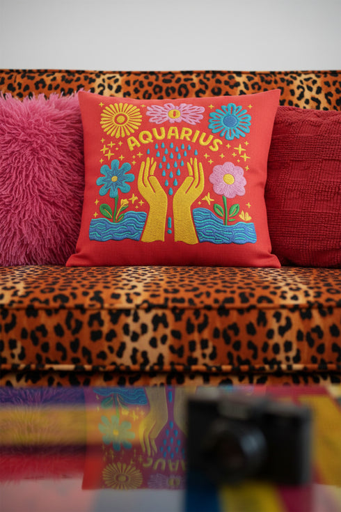A square red decorative pillow featuring a flat, faux-embroidery design of the Aquarius zodiac sign. The word "AQUARIUS" curves in yellow letters above two yellow hands catching falling blue raindrops. Stylized pink and blue folk-art flowers and green leaves flank the hands, with blue waves at the bottom. The design mimics the texture of stitched thread but is a smooth 2D print.