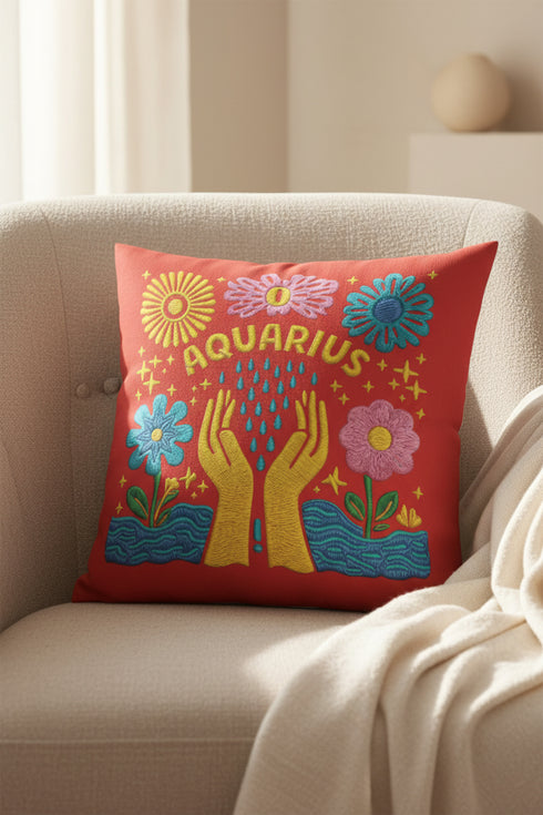A square red decorative pillow featuring a flat, faux-embroidery design of the Aquarius zodiac sign. The word "AQUARIUS" curves in yellow letters above two yellow hands catching falling blue raindrops. Stylized pink and blue folk-art flowers and green leaves flank the hands, with blue waves at the bottom. The design mimics the texture of stitched thread but is a smooth 2D print.
