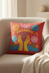 A square red decorative pillow featuring a flat, faux-embroidery design of the Aquarius zodiac sign. The word "AQUARIUS" curves in yellow letters above two yellow hands catching falling blue raindrops. Stylized pink and blue folk-art flowers and green leaves flank the hands, with blue waves at the bottom. The design mimics the texture of stitched thread but is a smooth 2D print.