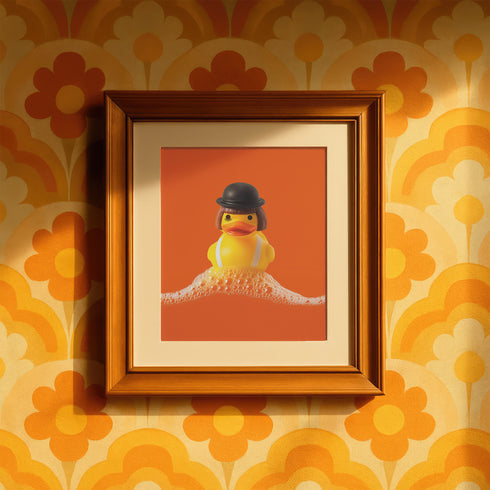 A fine art print of a rubber duck dressed like Alex from A Clockwork Orange, featuring a black bowler hat, blunt bob, and white suspenders. The duck floats in orange-tinged bubbles and is framed in white on a glossy pink tile bathroom shelf.