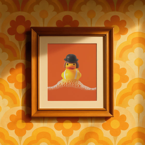 A fine art print of a rubber duck dressed like Alex from A Clockwork Orange, featuring a black bowler hat, blunt bob, and white suspenders. The duck floats in orange-tinged bubbles and is framed in white on a glossy pink tile bathroom shelf.
