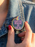 A round metal pinback button badge featuring a black background with the words QUEER MAGIC IS REAL in capital pink retro serif typography. A bright rainbow colored lightning bolt strikes from the top center, surrounded by concentric rainbow rings in red, orange, yellow, green, blue, and purple. The design is accented with small pink four-point stars and red crescent moons. The artwork features a realistic 2-D faux embroidery texture. The back has a silver metal safety pin clasp fastening.