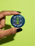 A round 1.75 inch pinback button featuring a retro psychedelic design on a dark navy blue background. The text reads BE KIND STAY WEIRD in vintage cream lettering around the border. The center illustration depicts a neon green skull stacked on top of a neon green alien head. Both the skull and alien have opening third eyes on their foreheads. Pink wavy lines frame the faces. The background is decorated with small colorful mushrooms, hearts, stars, and clouds. Glossy finish.