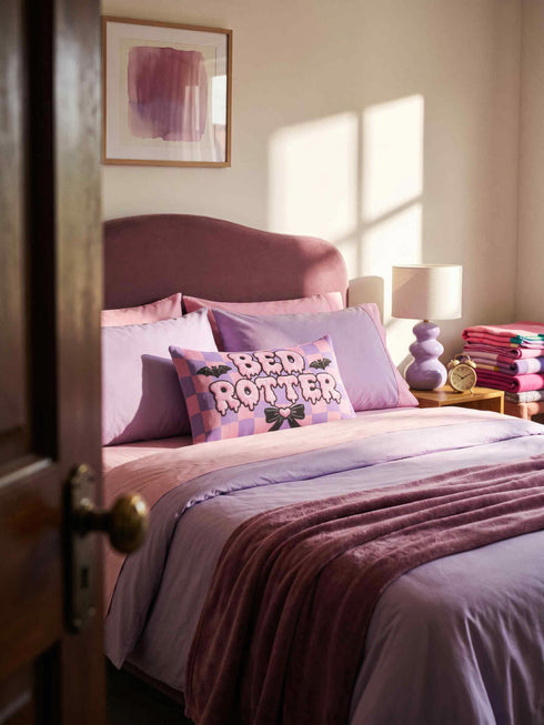 Pastel goth lumbar pillow, 20 by 12 inches, with a pink and lavender purple wavy checkerboard background. The words Bed Rotter appear in dripping pink bubble letters with a faux embroidery thread texture and black outlines. Two black bats with pink stitch details flank the text, and a black bow with a pink heart center sits below. Double sided flat print that mimics raised embroidery on linen feel polyester with a hidden zipper, insert included.