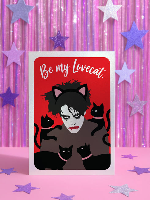 A vertical greeting card featuring a flat vector illustration of Robert Smith from The Cure against a vibrant red background. He has messy black hair, pale skin with smeared lipstick, and wears black cat ears. He is surrounded by four black cats with pink collars. White script text at the top reads "Be my Lovecat." The card is printed on shimmery pearlescent cardstock that catches the light. A high-quality alternative Valentine or anniversary card for goth music lovers and post-punk fans.
