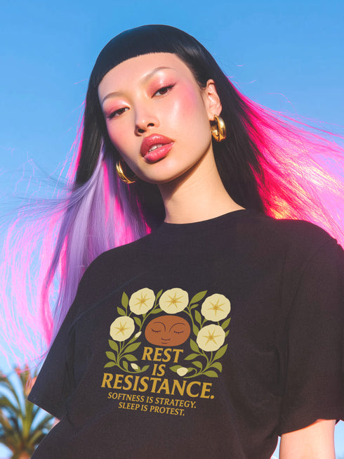 A black unisex t-shirt with a central front graphic. The design features the words "REST IS RESISTANCE" in a bold, gold, retro font. Above the main text, a serene, stylized face with closed eyes is framed by six cream-colored morning glory flowers and green leaves in a folk art, botanical style. Below, smaller text reads "SOFTNESS IS STRATEGY. SLEEP IS PROTEST." This alternative fashion, activist graphic tee is