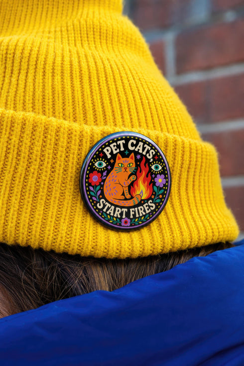 Glossy mylar pinback button featuring a solid black background. The central illustration shows a speckled orange cat sitting beside a stylized yellow and red flame. Bold retro text reads PET CATS, curved at the top, and START FIRES curved at the bottom. The design is framed by a colorful folk art border containing purple and red flowers, green leaves, and two floating eyes with light blue irises. The physical button is perfectly round with a highly reflective surface and a sturdy pin back.