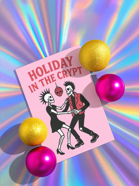 A 5x7 inch greeting card with a vibrant pink background and red retro text reading 'HOLIDAY IN THE CRYPT'. The illustration shows a male and female punk rock skeleton dancing under a red skull disco ball. The female skeleton has a mohawk and wears a black dress. The male skeleton has a mohawk and wears a black jacket with a red vest. Inside, the message reads, 'From our crypt to yours. Eat, drink, and be scary.' The style is a graphic, punk aesthetic.