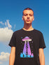 A person wearing a black unisex graphic t-shirt. The front of the shirt features a large, vibrant design with a retro-futuristic aesthetic. A classic flying saucer UFO, rendered in a pink and purple vaporwave gradient, hovers in the center. A beam of light descends from the UFO, containing the word 'RESIST' in a bold, uppercase font that transitions from purple to blue. Below the UFO, a stylized forest of neon blue and pink pine trees sits at the base of the design.