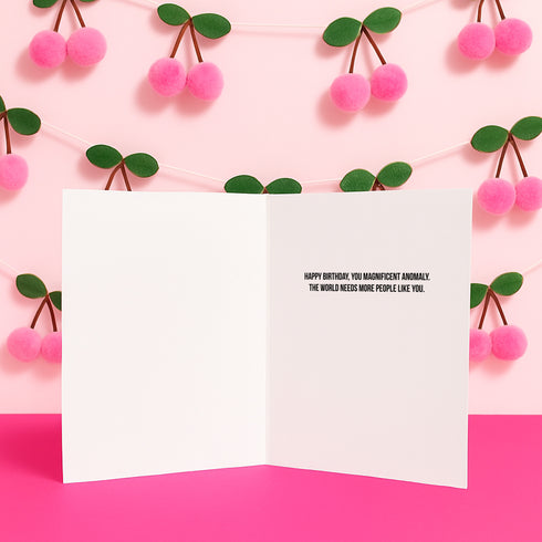 a birthday card with a message on it, placed against a pink background with pink pom-poms hanging from a string.