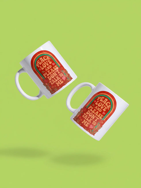 A glossy white porcelain ceramic mug featuring a vibrant maximalist design centered on the exterior. The artwork displays a retro 1970s-inspired rainbow arch composed of concentric bands in burnt orange, mustard yellow, and teal green. Inside the arch, stylized vintage bubble-serif typography in reddish-orange reads PEACE LOVE AND PLEASE DON'T TALK TO ME. The design evokes a groovy, hippie aesthetic mixed with sassy introvert humor.