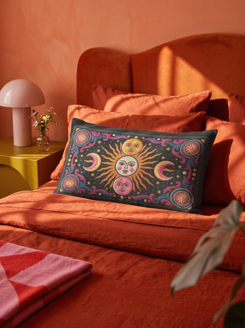 Rectangular 20x12 inch lumbar pillow cover in black featuring printed faux crewel embroidery of a large orange sunburst with a cream face, flanked by pink and yellow crescent moons, a golden moon face above and a pink moon face below, gold stars, blue dots, magenta and blue baroque scroll flourishes, and four corner medallions with golden flowers. Whimsigoth celestial design printed on both sides with a hidden zipper, crafted from soft linen look polyester.