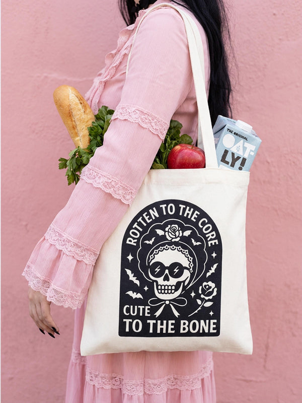 A flat rectangular canvas tote bag featuring a central gothic graphic on a natural cream background. The illustration depicts a detailed white human skull wearing retro sunglasses with lightning bolts and a scalloped bonnet with roses. The skull is framed by a black archway with silhouette bats and stars. Text reads "Rotten to the Core" and "Cute to the Bone." High contrast black and white colors pop against the fabric, perfect for whimsigoth fashion.