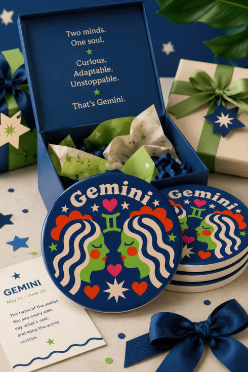 A close-up photograph of a round, glossy ceramic coaster featuring a bold retro pop art design. The background is a deep royal blue. In the center are two symmetrical profile faces of twins looking at each other, featuring bright green skin, closed eyes, and red cheeks. They have wavy white and pale pink hair with red accents. The word Gemini is arched at the top in a retro white font. The design is decorated with a green Gemini symbol, pink and red hearts, and white starbursts.
