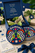 A close-up photograph of a round, glossy ceramic coaster featuring a bold retro pop art design. The background is a deep royal blue. In the center are two symmetrical profile faces of twins looking at each other, featuring bright green skin, closed eyes, and red cheeks. They have wavy white and pale pink hair with red accents. The word Gemini is arched at the top in a retro white font. The design is decorated with a green Gemini symbol, pink and red hearts, and white starbursts.