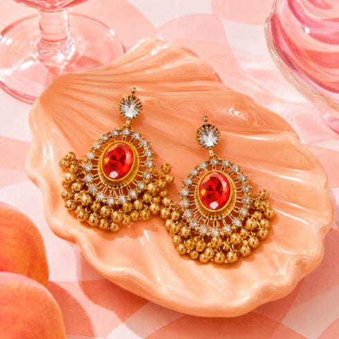 A pair of ornate earrings with a large red gemstone and gold beads are displayed on a pink shell-shaped plate, accompanied by a glass of wine and peaches.