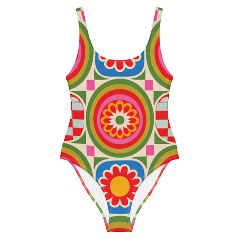 A one-piece swimsuit with a 1970s retro psychedelic geometric pattern in red, orange, green, and blue on a cream background. The swimsuit features a scoop neck and cheeky high-cut fit. In one view, she styles it with a pink baseball cap and gold necklaces. The design includes a central radial sunburst mandala and checkerboard side panels, representing a maximalist, rockstar-ready aesthetic.