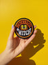 A round, black embroidered patch with a thick, textured merrow border stitched in alternating orange and yellow thread. In the center, the words "PAPERBACK WITCH" are embroidered in a retro 70s-style font. The word "PAPERBACK" is arched across the top in bold orange thread with a thin yellow outline. The word "WITCH" is across the bottom in puffy, bright white thread with a thin yellow outline. Between the words is a central icon of an open book, containing a stylized all-seeing eye.