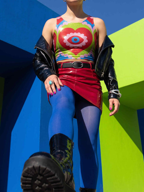 A sleeveless scoop neck crop top featuring a bold all-over printed design. The central graphic is a large bright red heart enclosing a scalloped pink flower shape and a blue evil eye motif in the middle. The background surrounds the heart with mirrored concentric wavy arches and scalloped borders in vibrant shades of lime green, pink, blue, and red. The fitted garment has a smooth athletic texture. Available in a standard matte performance knit or a sparkly silver glitter foil finish.