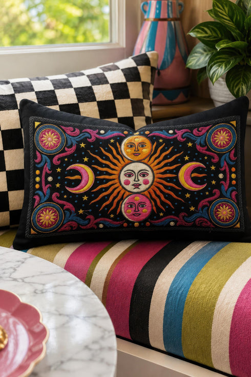 Rectangular 20x12 inch lumbar pillow cover in black featuring printed faux crewel embroidery of a large orange sunburst with a cream face, flanked by pink and yellow crescent moons, a golden moon face above and a pink moon face below, gold stars, blue dots, magenta and blue baroque scroll flourishes, and four corner medallions with golden flowers. Whimsigoth celestial design printed on both sides with a hidden zipper, crafted from soft linen look polyester.