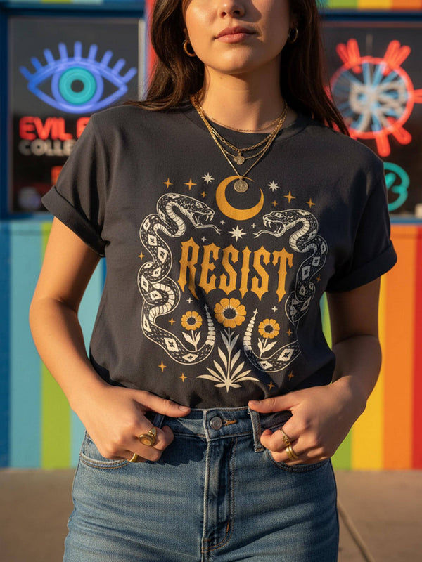 A unisex short sleeve graphic t shirt in dark charcoal black featuring a highly detailed symmetrical medallion print on the chest. In the center, the word RESIST is printed in a bold yellow gothic blackletter font. Two coiled white snakes with fine black scale details frame the text on the left and right. Above the text, a yellow crescent moon is flanked by white and yellow starbursts. Beneath, three yellow marigold flowers sit atop white stems and leafy foliage.