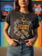 A unisex short sleeve graphic t shirt in dark charcoal black featuring a highly detailed symmetrical medallion print on the chest. In the center, the word RESIST is printed in a bold yellow gothic blackletter font. Two coiled white snakes with fine black scale details frame the text on the left and right. Above the text, a yellow crescent moon is flanked by white and yellow starbursts. Beneath, three yellow marigold flowers sit atop white stems and leafy foliage.