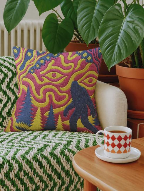 A square decorative throw pillow cover featuring a trippy retro Bigfoot scene. A large navy blue silhouette of Sasquatch walks through a forest of navy and yellow pine trees. The background is a psychedelic swirling sky in vibrant pink and yellow, featuring a large stylized eye and a cluster of small yellow stars. The flat two dimensional print visually mimics the textured look of heavy needlepoint embroidery. The pillow features a sleek hidden zipper on the bottom. No insert is included.