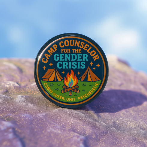Round glossy pinback button measuring 2.25 inches featuring a playful camp-themed design with the phrase “Camp Counselor for the Gender Crisis.” The artwork includes two orange tents, a rainbow campfire, stars, and green grass. Bottom text reads “Cabin Queer, Unit Psychedelic.” Made from durable tinplate with a safety pin back and coated in a UV and scratch-resistant mylar finish. Bold and colorful, perfect for jackets, bags, or pride outfits.