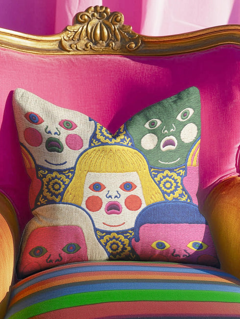 A square decorative pillow cover featuring a faux embroidery pattern of five stylized, expressive doll faces. The central face has yellow hair and pink cheeks. It is surrounded by four other faces in shades of pale beige, green, and bright pink, all with wide staring eyes and open mouths. The design uses intricate, colorful line work to create the illusion of thick woven stitching, but the fabric is a smooth print. Medallions fill the gaps.