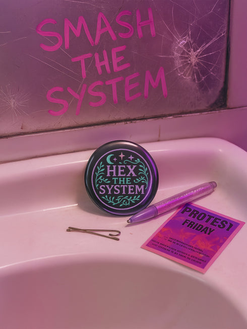 Glossy black pinback button with pastel embroidery-style design that says “Hex the System,” surrounded by vines, stars, and a crescent moon. Displayed against witchy pink and blue backdrops, street pole flyers, and protest props.