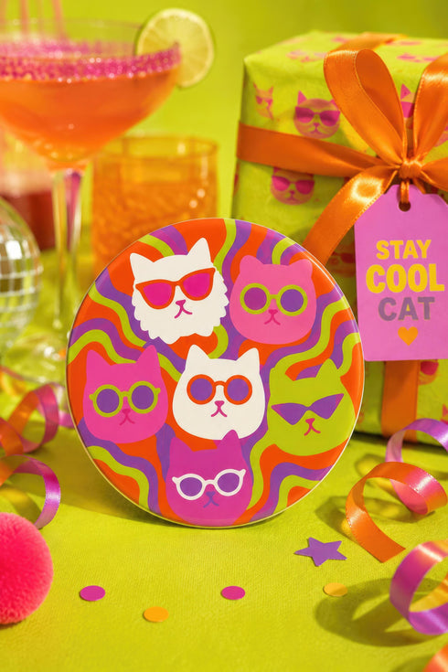 A close-up photograph of a glossy round ceramic coaster featuring a retro 70s psychedelic design. The artwork displays a cluster of six stylized cat face illustrations, each wearing a different pair of sunglasses including retro round and cat-eye shapes. The cats are colored in solid shades of white, hot pink, purple, and lime green. They are set against a vibrant background of wavy, swirling lines in bright orange, red, and purple. The top surface has a highly reflective clear glaze.
