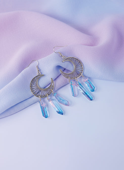 A pair of earrings with a crescent moon design and crystal pendants, resting on a purple fabric background.